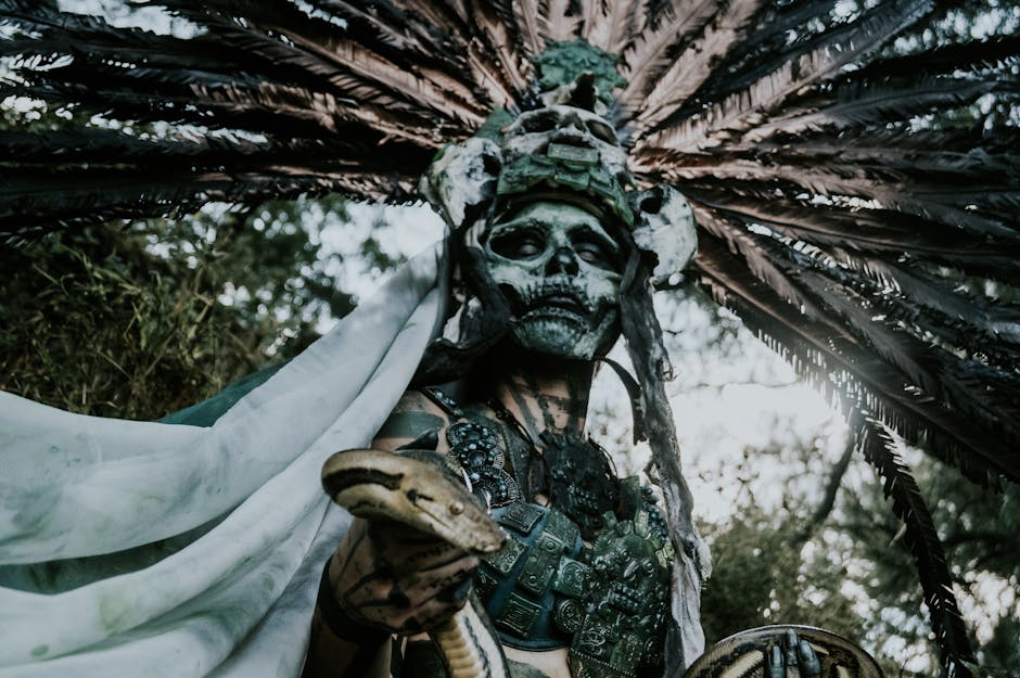 A mysterious figure adorned in a feathered headdress and skull mask holds a snake outdoors.